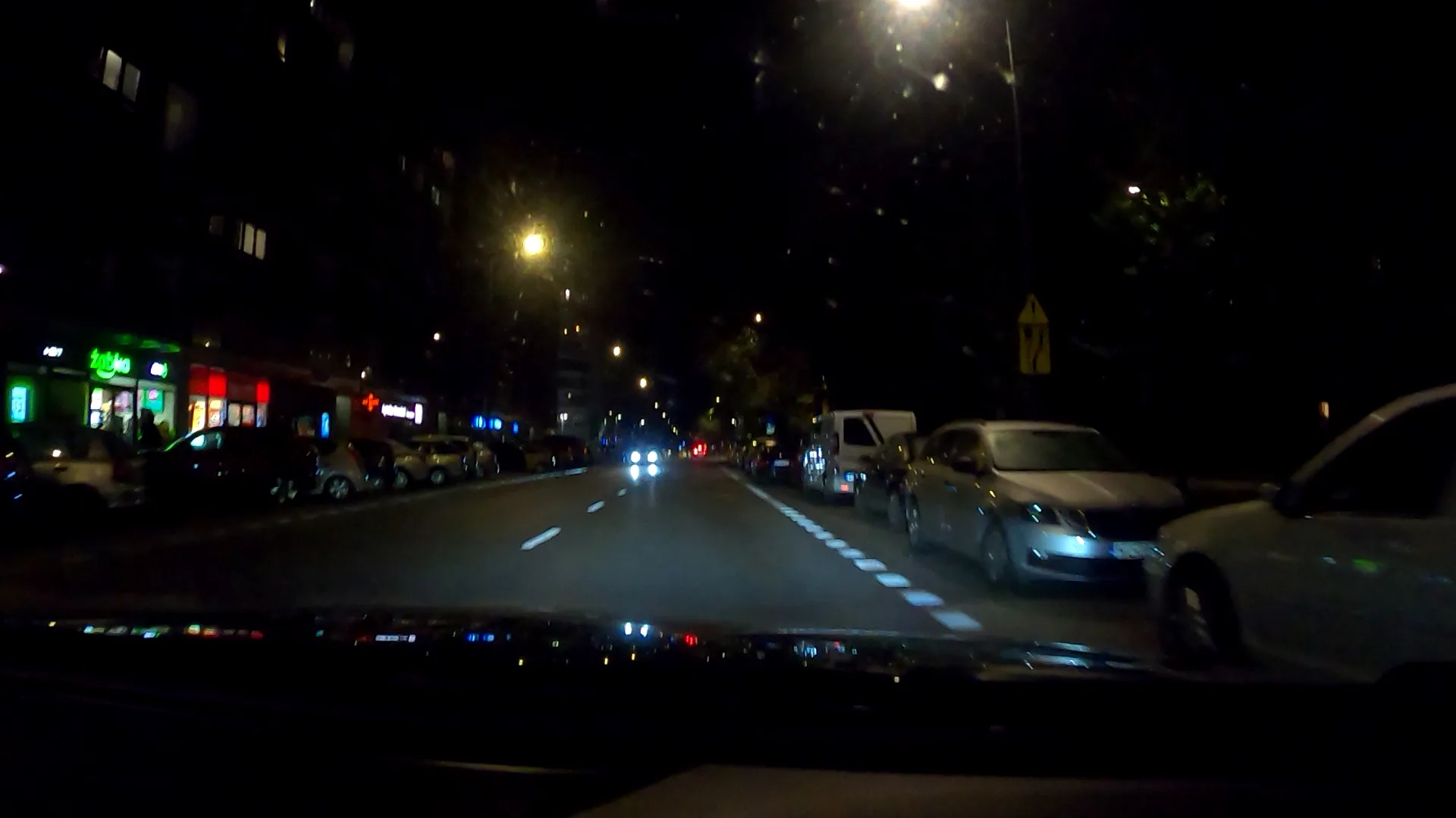 Night street recording through a vehicle window; the signs of parked cars are small and blurry